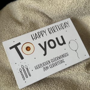 Op de afbeelding: Een witte verjaardagskaart met zwarte tekst "HAPPY BIRTHDAY To you". De kaart bevat illustraties van een ballon, kaarsen en een donut. De Duitse zin "HERZLICHEN GLÜCKWUNSCH ZUM GEBURTSTAG" staat er ook op.