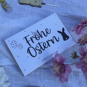 Peut inclure: Une carte blanche avec l'inscription "Frohe Ostern" et une illustration de lapin, ainsi que des œufs de Pâques décoratifs. La carte est accompagnée de friandises de Pâques au chocolat et de fleurs roses.