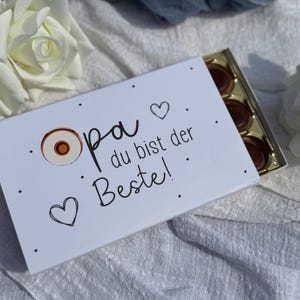 Peut inclure: Une boîte cadeau blanche avec le texte allemand "Opa du bist der Beste!" (Grandpa, tu es le meilleur !) est ouverte, révélant plusieurs chocolats. La boîte est décorée de cœurs et de points.