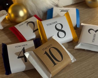 Pode incluir: Close-up de um calendário do advento com pacotes numerados em várias cores. Os pacotes são embrulhados em papel branco e têm números impressos. Um gnomo decorativo e ornamentos dourados no fundo.