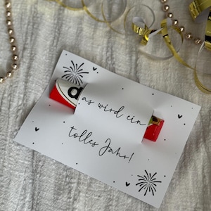 May include: A white card with the text "as wird ein tolles Jahr!" and black fireworks and heart illustrations. A red and white candy bar is partially visible. Gold ribbon and beads are in the background.