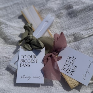 Könnte beinhalten: Eine Ansammlung weißer Papierfächer mit Anhängern. Auf den Anhängern steht "TO OUR BIGGEST FANS" und "stay cool". Die Fächer sind mit olivgrünen und altrosa Bändern gebunden, was eine weiche, elegante Ästhetik erzeugt.