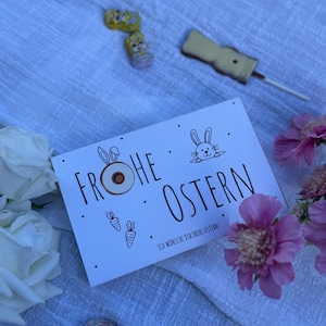 Puede incluir: Una tarjeta de felicitación de Pascua con la frase "Frohe Ostern" y una ilustración de un conejito. La tarjeta está rodeada de bombones, una piruleta y flores rosas, todo dispuesto sobre un paño blanco.