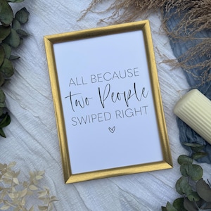 May include: A framed print with a gold-coloured frame. The white print features the text "ALL BECAUSE TWO PEOPLE SWIPED RIGHT" in black script, with a small heart symbol below. The frame is rectangular.