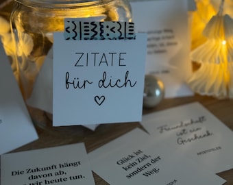 Puede incluir: Un tarro de cristal con tapa, lleno de pequeñas tarjetas blancas con citas en alemán. El tarro está decorado con una cinta adhesiva estampada. Una pequeña casa blanca y luces de hadas son visibles en el fondo.