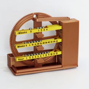 May include: A copper-coloured mechanical clock with a large gear and three yellow tracks labelled "Minute" and "Hour." Small metal balls move along the tracks to indicate the time. The clock's design is intricate and features a rectangular base.