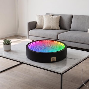 May include: A circular black kinetic sand table featuring a glowing LED ring that illuminates the sand surface with a vibrant rainbow gradient. The device has a small square QR code label on the side and a white power cord extending from the base.