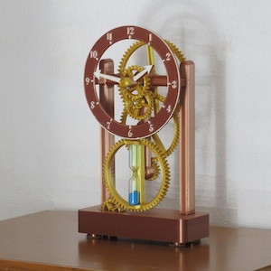 May include: A mechanical clock featuring a brown circular dial with white numbers and white hands. The structure includes gold-coloured gears and a central hourglass containing blue sand, all supported by a brown rectangular base and vertical frame.