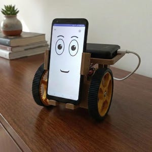 May include: A smartphone with a cartoon face displayed on the screen is mounted on a wooden cart with yellow wheels and black tires. A black device sits on top of the cart, connected by a white cable.