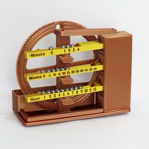 May include: A copper-colored mechanical clock with a large gear and three yellow tracks labeled "Minute" and "Hour." Small silver balls indicate the time. The clock's design is intricate and appears to be 3D-printed.