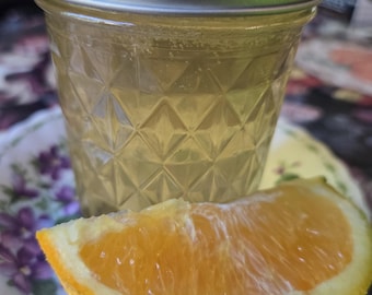 May include: A clear glass jar with a diamond-patterned texture and a silver metal lid, filled with a pale yellow liquid. A fresh, bright orange citrus wedge is positioned directly in front of the glass jar, resting on a floral patterned plate.