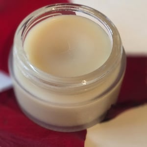 May include: A small, clear glass jar filled with a creamy, off-white substance. The jar sits on a vibrant red rose petal, with a white background. The product appears to be a cosmetic item, such as a balm or cream.