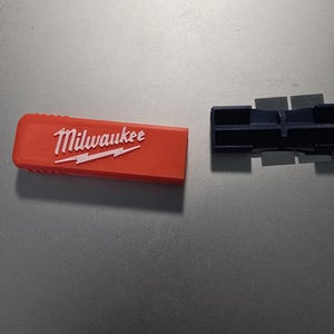 May include: An orange rectangular tool accessory with the white "Milwaukee" logo and lightning bolt design. A dark blue tool component is next to it. The items are on a grey surface.