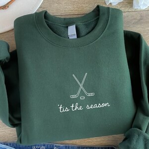 May include: A dark green sweatshirt with white text and a hockey-themed design. The text reads "'tis the season" below a graphic of crossed hockey sticks and a puck. The sweatshirt is folded on a wooden surface.