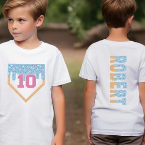 May include: White t-shirt with a blue and white ice cream cone design on the front. The design includes the number 10 and a baseball diamond. The back of the shirt has the name "ROBERT" spelled vertically in a similar ice cream cone design.
