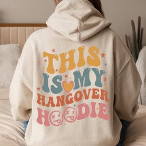 May include: A beige hoodie with the text "THIS IS MY HANGOVER HOODIE" in retro-style lettering. The design includes stars, a heart, and smiley faces. The hoodie is worn by a person sitting on a bed.