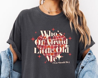 May include: A dark gray t-shirt with a red, white, and gold graphic of a disco ball with the text "Who's Afraid Little Old Me (You Should Be)"