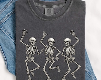 May include: Dark gray t-shirt featuring a graphic of three dancing skeletons. The skeletons are in a playful pose with their arms raised. The t-shirt is laid on top of a pair of blue denim jeans.