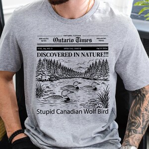 May include: A gray t-shirt featuring a newspaper-style graphic with the headline "DISCOVERED IN NATURE!!!" and the text "Stupid Canadian Wolf Bird." The graphic includes a lake scene with loons and trees. The shirt also has the text "Ontario Times."