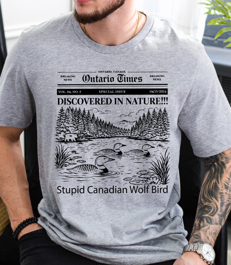 May include: A gray t-shirt featuring a newspaper-style graphic with the headline "DISCOVERED IN NATURE!!!" and the text "Stupid Canadian Wolf Bird." The graphic includes a lake scene with loons and trees. The shirt also has the text "Ontario Times."