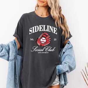 Op de afbeelding: Donkergrijs t-shirt met de tekst "SIDELINE SOCIAL CLUB" en een honkbalhandschoenafbeelding. De afbeelding bevat een rode handschoen en een witte honkbal. Het shirt heeft een vintage, versleten uitstraling.