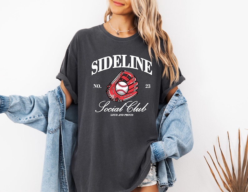 Op de afbeelding: Donkergrijs t-shirt met de tekst "SIDELINE SOCIAL CLUB" en een honkbalhandschoenafbeelding. De afbeelding bevat een rode handschoen en een witte honkbal. Het shirt heeft een vintage, versleten uitstraling.