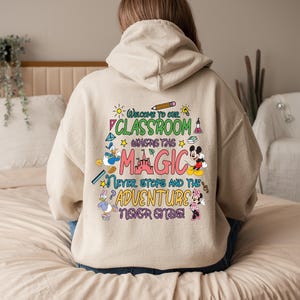 May include: A beige hoodie with a colourful graphic print that says "Welcome to our classroom where the magic never stops and the adventure never ends". The graphic includes Mickey Mouse, Donald Duck, and other Disney characters.
