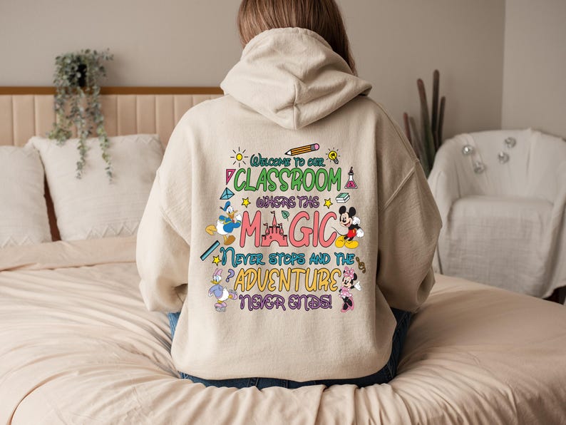 May include: A beige hoodie with a colourful graphic print that says "Welcome to our classroom where the magic never stops and the adventure never ends". The graphic includes Mickey Mouse, Donald Duck, and other Disney characters.