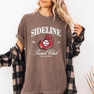 Op de afbeelding: Een bruin T-shirt met de tekst "SIDELINE SOCIAL CLUB" en een honkbalhandschoenafbeelding. De afbeelding bevat een honkbal en het nummer 23. Het shirt heeft een casual stijl.
