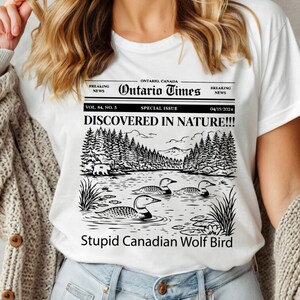 May include: White t-shirt featuring a newspaper-style graphic with the headline "DISCOVERED IN NATURE!!!" and the text "Stupid Canadian Wolf Bird." The graphic includes a lake scene with loons and a forest. The shirt also has the text "Ontario Times."