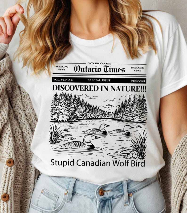 May include: White t-shirt featuring a newspaper-style graphic with the headline "DISCOVERED IN NATURE!!!" and the text "Stupid Canadian Wolf Bird." The graphic includes a lake scene with loons and a forest. The shirt also has the text "Ontario Times."