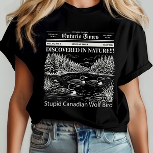 May include: Black t-shirt featuring a newspaper-style graphic with the headline "DISCOVERED IN NATURE!!!" and the text "Stupid Canadian Wolf Bird." The graphic includes a lake scene with loons and a forest.