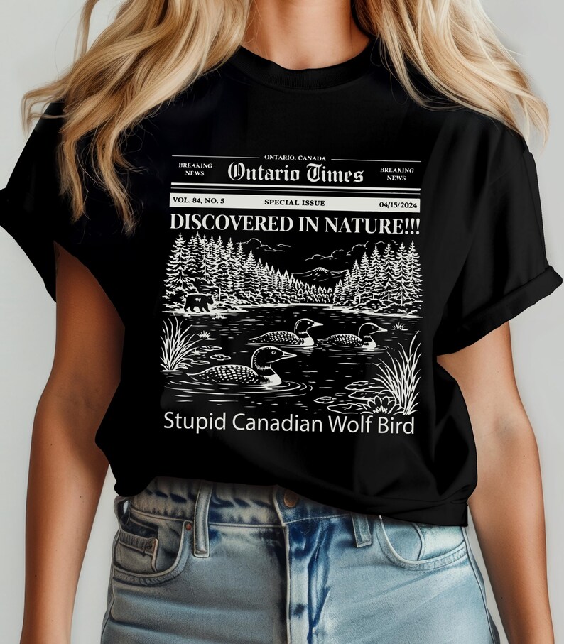 May include: Black t-shirt featuring a newspaper-style graphic with the headline "DISCOVERED IN NATURE!!!" and the text "Stupid Canadian Wolf Bird." The graphic includes a lake scene with loons and a forest.