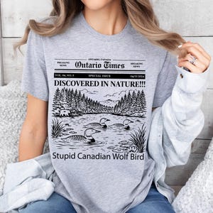 May include: A light gray t-shirt featuring a black and white graphic of a lake scene with ducks, trees, and the text "DISCOVERED IN NATURE!!!" and "Stupid Canadian Wolf Bird". The shirt also has the text "Ontario Times".