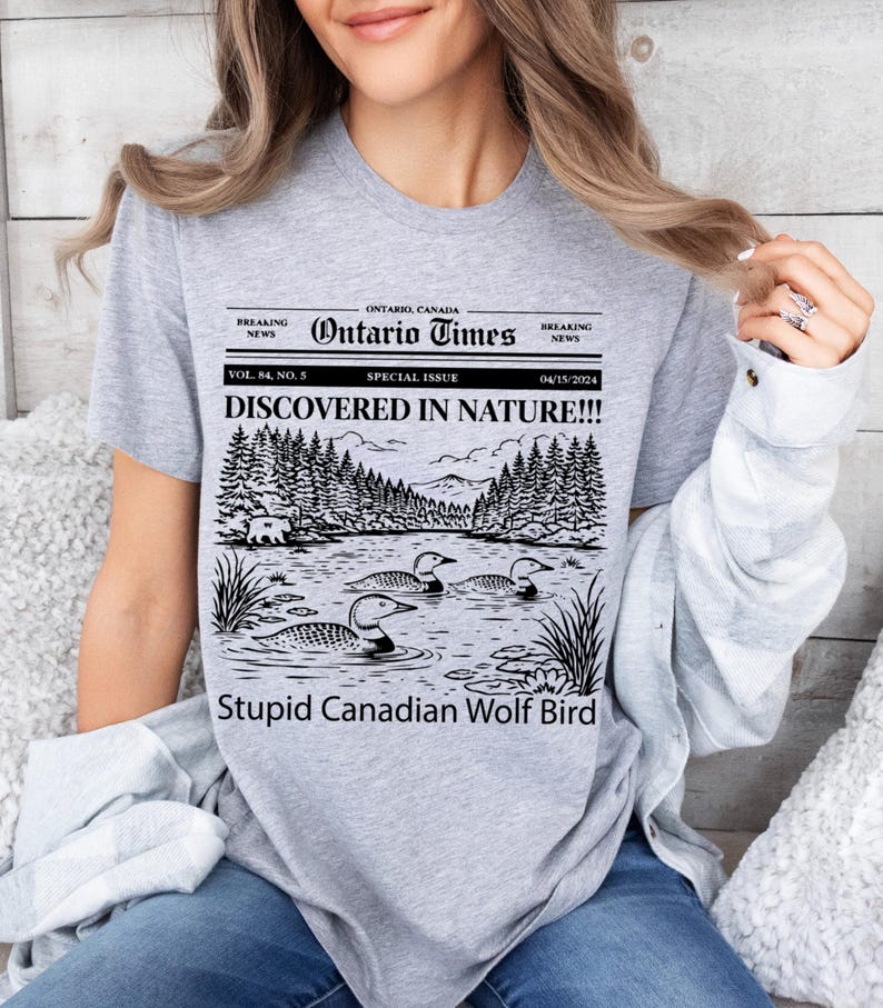 May include: A light gray t-shirt featuring a black and white graphic of a lake scene with ducks, trees, and the text "DISCOVERED IN NATURE!!!" and "Stupid Canadian Wolf Bird". The shirt also has the text "Ontario Times".