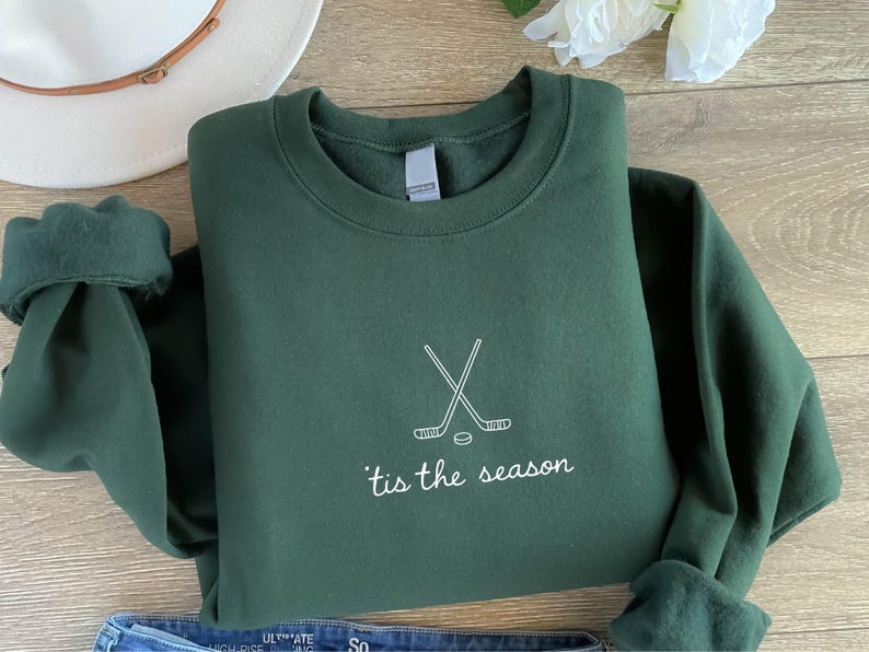 May include: A dark green sweatshirt with white text and a hockey-themed design. The text reads "'tis the season" below a graphic of crossed hockey sticks and a puck. The sweatshirt is folded on a wooden surface.