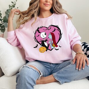 May include: A light pink sweatshirt featuring a heart-shaped design with Jack Skellington and Sally from The Nightmare Before Christmas. The design includes bats and a carved pumpkin. The sweatshirt is a crew neck style.