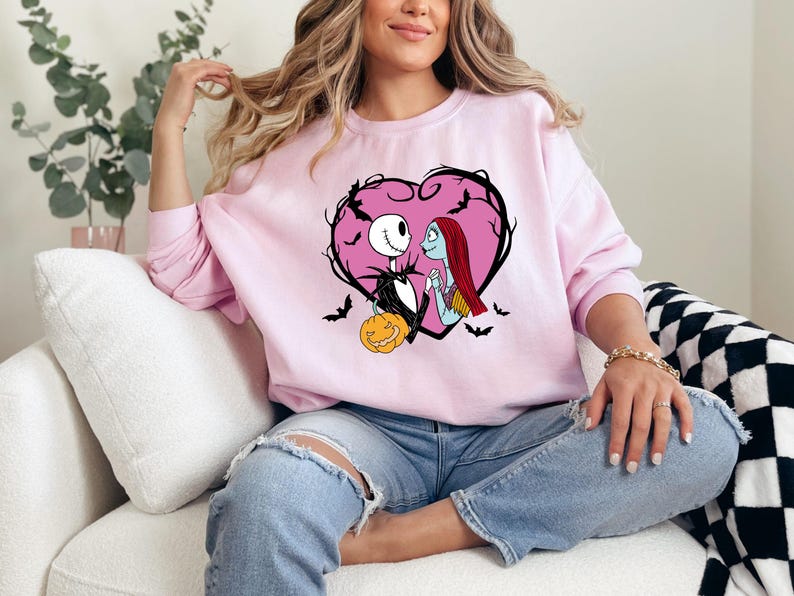 May include: A light pink sweatshirt featuring a heart-shaped design with Jack Skellington and Sally from The Nightmare Before Christmas. The design includes bats and a carved pumpkin. The sweatshirt is a crew neck style.