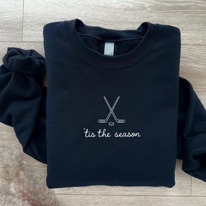 May include: A navy blue sweatshirt with a crew neck. The front features a white embroidered design of crossed hockey sticks and a puck, with the phrase "'tis the season" below. The sweatshirt is folded on a light wood surface.