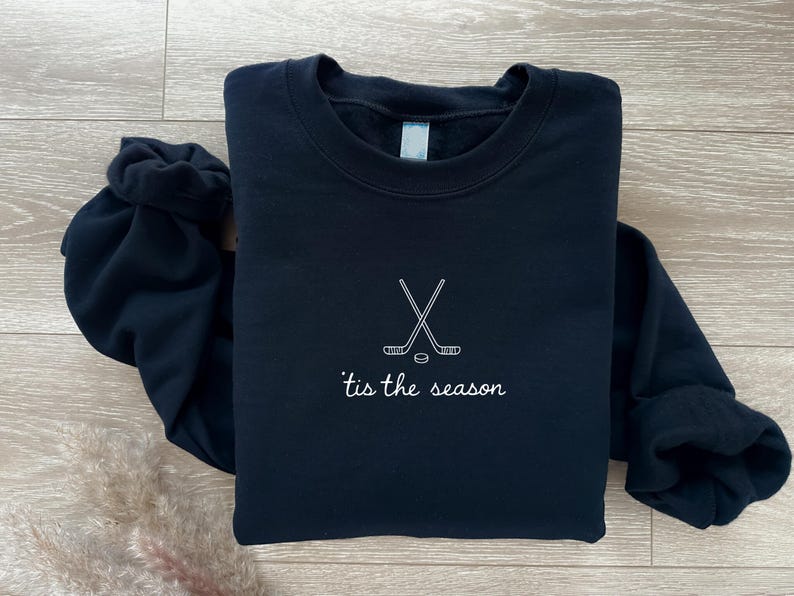 May include: A navy blue sweatshirt with a crew neck. The front features a white embroidered design of crossed hockey sticks and a puck, with the phrase "'tis the season" below. The sweatshirt is folded on a light wood surface.
