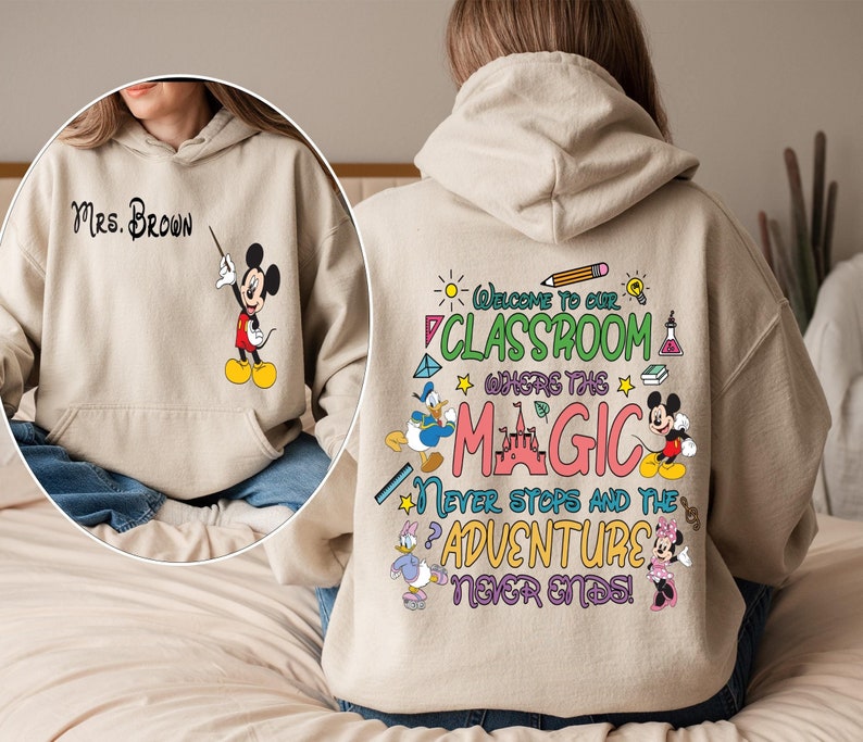 May include: Beige hoodie with a Mickey Mouse design on the front and a colourful graphic on the back that says "Welcome to our classroom where the magic never stops and the adventure never ends!"