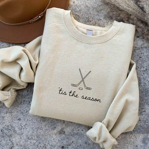 May include: A cream-colored sweatshirt with the text "'tis the season" and a hockey stick and puck graphic. The long-sleeved top is folded on a textured gray surface, with a brown hat visible in the background.
