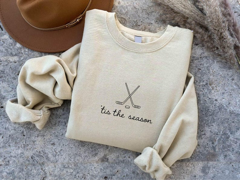 May include: A cream-colored sweatshirt with the text "'tis the season" and a hockey stick and puck graphic. The long-sleeved top is folded on a textured gray surface, with a brown hat visible in the background.
