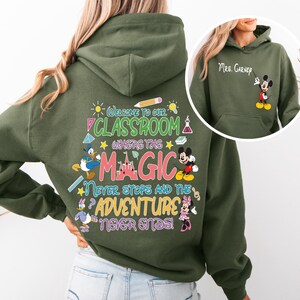 May include: Green hooded sweatshirt with a colourful graphic print featuring Mickey Mouse, Minnie Mouse, Donald Duck, and a castle. The text reads "Welcome to our classroom where the magic never stops and the adventure never ends!"