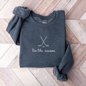 May include: A dark gray sweatshirt with white text and a hockey-themed design. The design features crossed hockey sticks and a puck, with the phrase "'tis the season" below. The sweatshirt is folded on a light wood-grain surface.