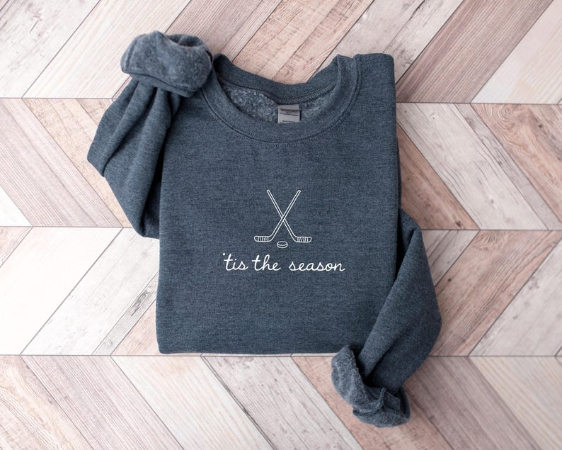 May include: A dark gray sweatshirt with white text and a hockey-themed design. The design features crossed hockey sticks and a puck, with the phrase "'tis the season" below. The sweatshirt is folded on a light wood-grain surface.