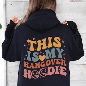 May include: A navy blue hoodie with the text "THIS IS MY HANGOVER HOODIE" in retro-style lettering. The design features orange, yellow, teal, and pink colors with star and heart accents. The hoodie has a hood and long sleeves.