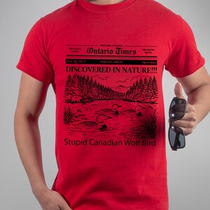 May include: Red t-shirt with a black graphic resembling a newspaper headline. The headline reads "Ontario Times" and "DISCOVERED IN NATURE!!!". The graphic includes a lake scene with birds and the text "Stupid Canadian Wolf Bird".