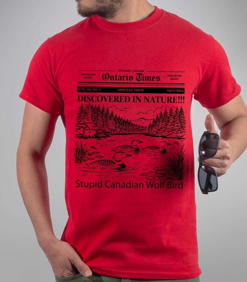 May include: Red t-shirt with a black graphic resembling a newspaper headline. The headline reads "Ontario Times" and "DISCOVERED IN NATURE!!!". The graphic includes a lake scene with birds and the text "Stupid Canadian Wolf Bird".