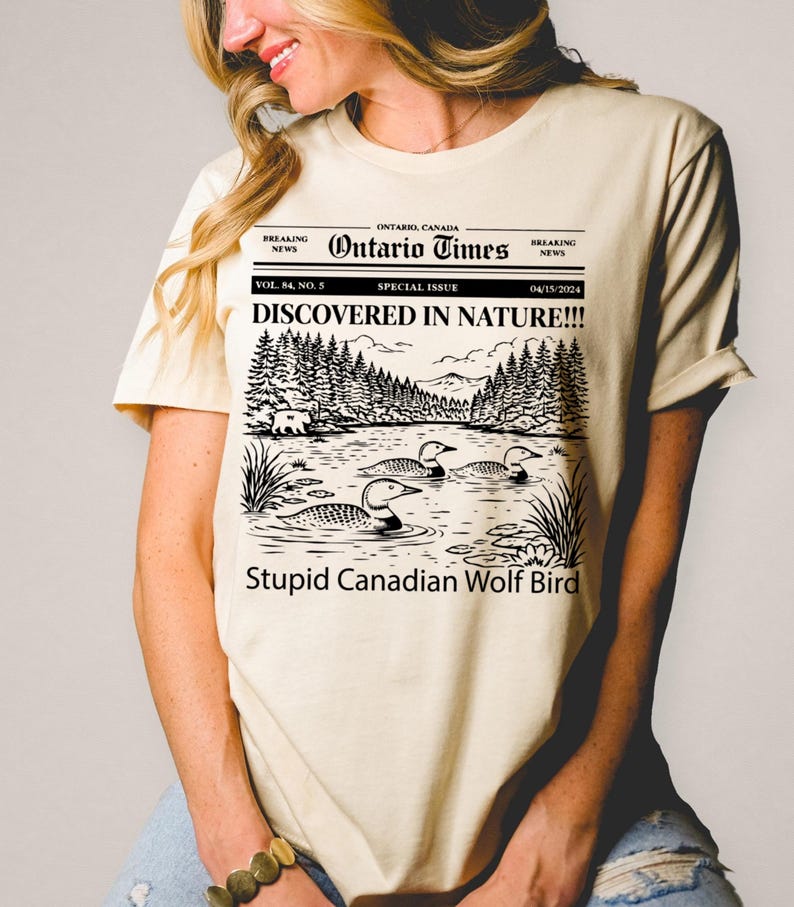 May include: Cream-colored t-shirt featuring a newspaper-style graphic with the headline "Ontario Times." The graphic includes a lake scene with loons and the text "DISCOVERED IN NATURE!!!" and "Stupid Canadian Wolf Bird."